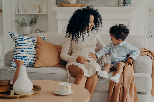 Lovely pregnant mixed race woman mother explaining to curious son about his future brother or sister, cute little boy touch belly of his mom and talking with baby while sitting on couch in living room