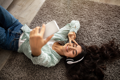 Above view of lovely Indian lady taking selfie while listening to music in headphones on floor at home. Charming millennial woman making photo of herself, enjoying her favorite song