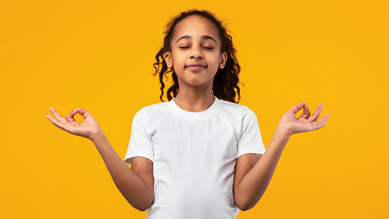 Zen, Mindfulness, Balance and Peace Concept. Portrait of calm black little girl meditating with closed eyes, holding her fingers in mudra gesture, yellow studio background, copyspace, banner, panorama