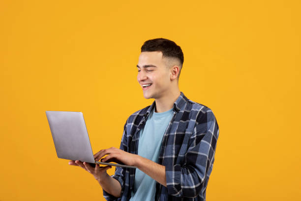 istock Happy young man looking at laptop screen with smile, watching interesting web content on orange studio background 1322848220