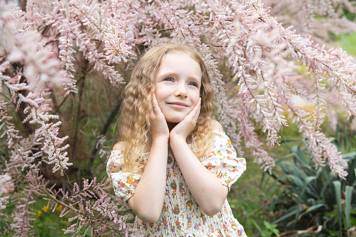 Portrait of a happy little girl with blond curly hair and in a dress. The child enjoys a walk in the botanical park.