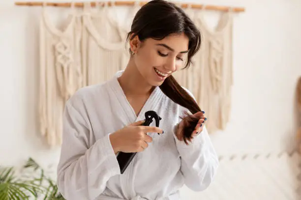 Split Ends Treatment. Brunette Lady Spraying Detangling Spray On Hair Moisturizing Dry Ends Standing In Modern Bathroom At Home, Wearing Bathrobe. Haircare Cosmetics And Beauty Routine Concept