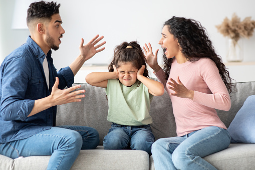 Parental Quarrels. Arab Man And Woman Arguing In Front Of Their Child, Upset Little Girl Covering Ears With Hands Not To Listen Conflict, Stressed Female Kid Sitting Between Shouting Parents