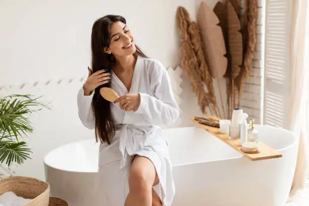 Haircare. Brunette Female Brushing Hair With Hairbrush Detangling Split Ends Smiling Sitting On Bathtub Wearing Bathrobe In Modern Bathroom At Home. Beauty Routine Concept