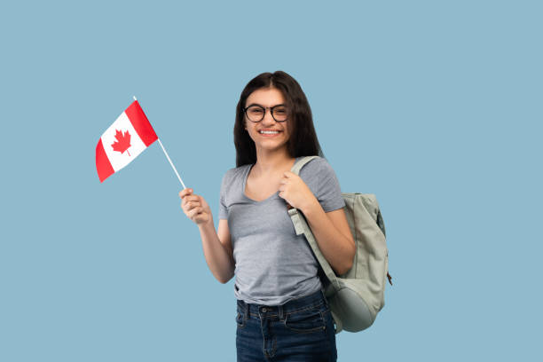 Happy Indian teen exchange student with backpack holding small flag of Canada and smiling at camera on blue background Happy Indian teen exchange student with backpack holding small flag of Canada and smiling at camera on blue studio background. Travel and modern foreign education concept Study Abroad Countries stock pictures, royalty-free photos & images