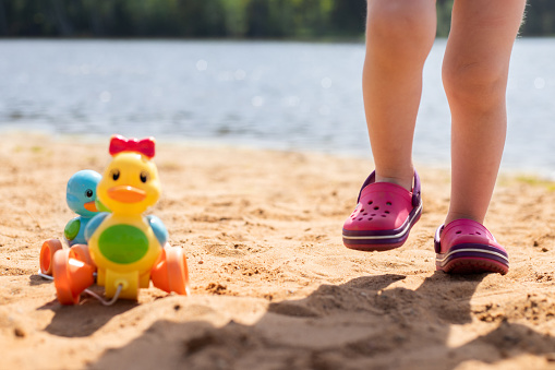 Children's feet in rubber pink slippers with a toy yellow duck on the beach on the water background