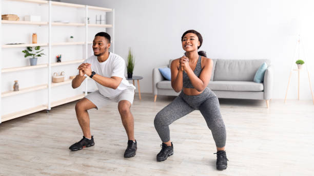 Strength workout concept. Fit black woman and her boyfriend doing squats together at home, panorama Strength workout concept. Fit black woman and her boyfriend doing squats together at home, panorama. African American couple having domestic training during covid-19 quarantine crouching stock pictures, royalty-free photos & images