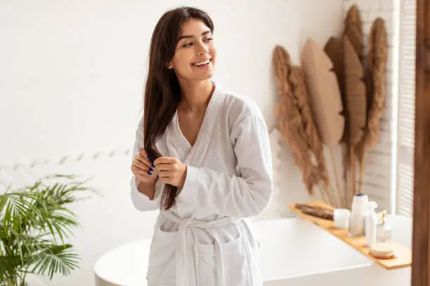 Photo of Joyful Woman In Bathrobe Touching Long Hair Standing In Bathroom