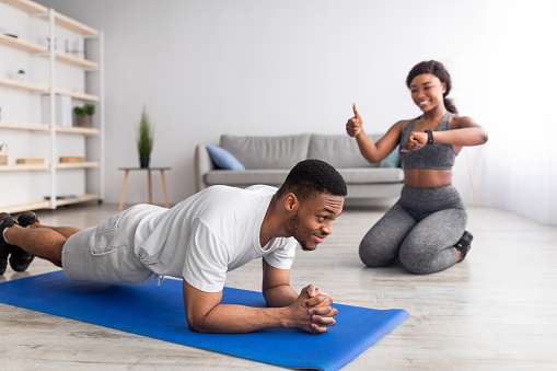 Fit black man standing in plank pose, his girlfriend noting time on smartwatch, showing thumb up gesture, supporting her boyfriend at home. African American couple training together as team