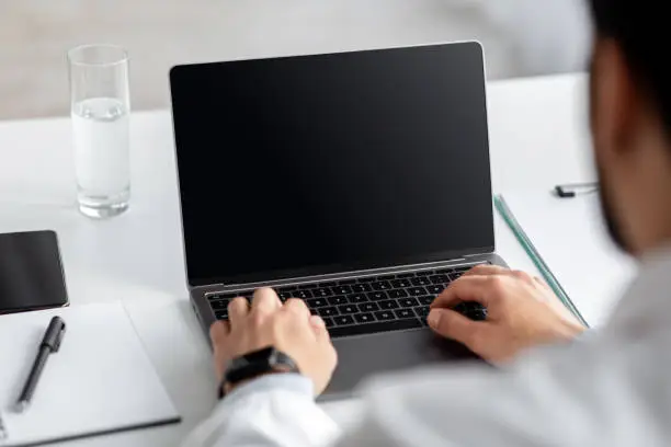 Photo of Millennial doctor or businessman in coat works on laptop with empty screen for text on white desk