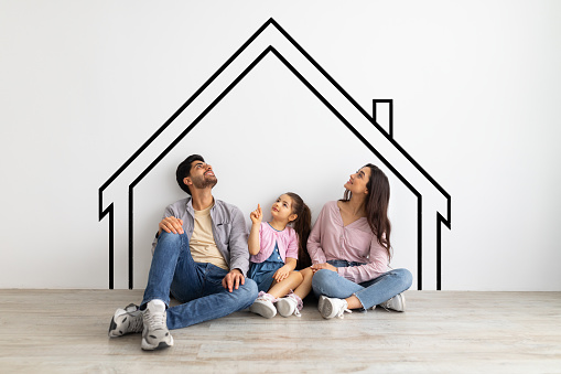 Relocation concept. Young eastern family sitting on floor and looking upwards, dreaming about something, leaning on white wall with painted house, collage with illustrations on white wall