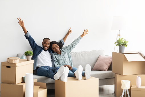 Happy Owners. Joyful Black Spouses Relaxing On Couch Among Boxes After Relocation, Cheerful Young African American Couple Celebrating Moving To New Apartment, Raising Hands With Joy, Copy Space