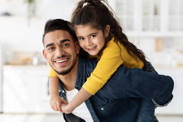 Arab Father And Daughter. Loving Young Man Carrying Little Girl On Back Arab Father And Daughter. Loving Young Man Carrying Little Girl On Back And Smiling At Camera , Cute Small Female Child Piggybacking Dad While They Having Fun At Home Together, Closeup Portrait arabic girl stock pictures, royalty-free photos & images