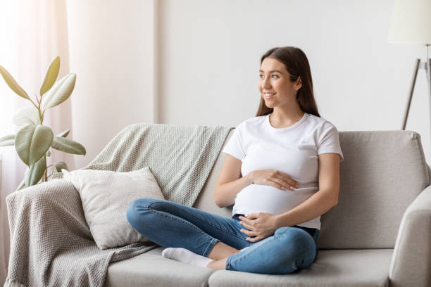 居心地の良いリビングルームでソファに座っている若い幸せな笑顔の妊婦 - abdomen adult affectionate baby ストックフォトと画像