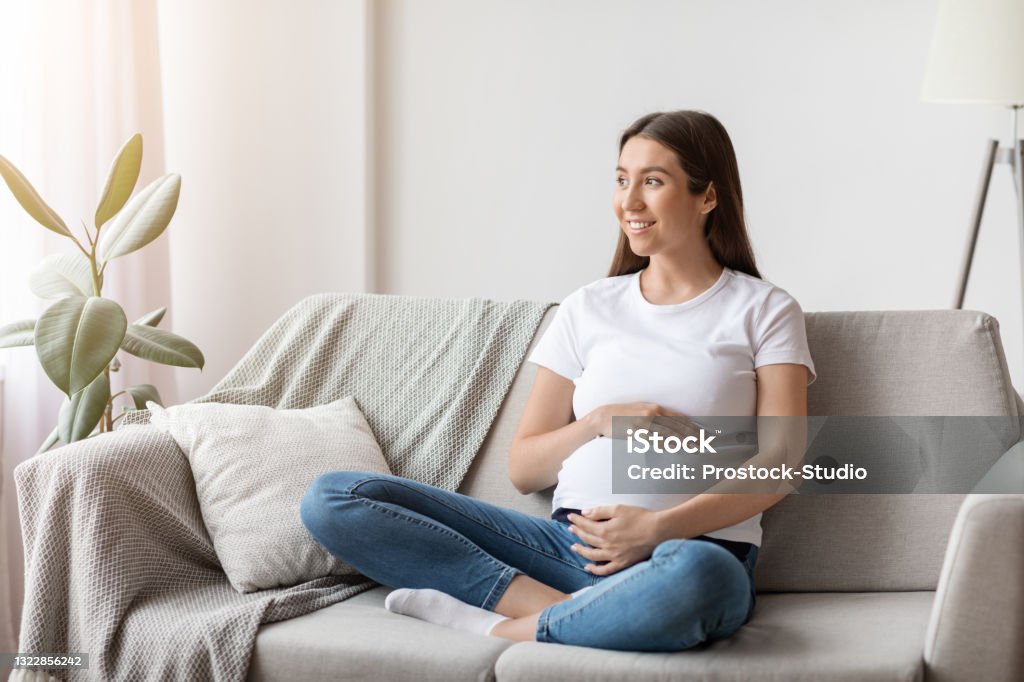 Young happy smiling pregnant woman sitting on couch in cozy living room Portrait of young happy smiling pregnant woman sitting on couch in cozy living room, beautiful expectant mother tenderly embracing belly and looking away, thinking about upcoming motherhood Pregnant Stock Photo