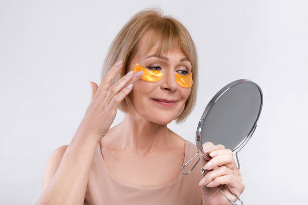 istock Sensual senior lady applying eye patches in front of mirror on light studio background 1322856334