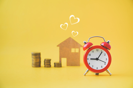 red analog clock ,blurred stack of coins and brown paper with heart shape on yellow background , new house, sweet home or resident , future planning for warm, perfect family concept