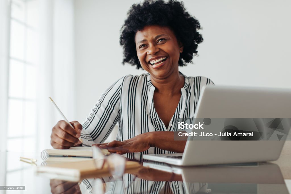 Happy mature woman working at home office Portrait of a smiling woman sitting at table with laptop and dairy. Woman smiling at camera while working from home office. Women Stock Photo