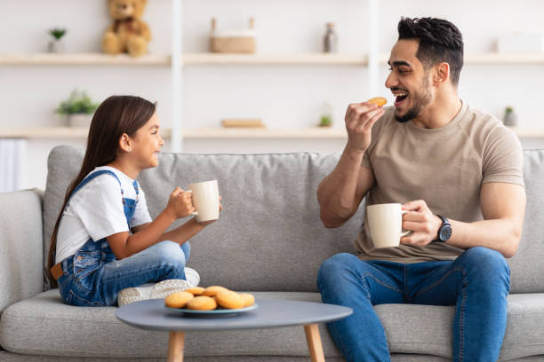 Cheerful little daughter and dad having breakfast Snack Time. Portrait of cheerful little girl and man holding cups, drinking coffee and tea, eating cookies and looking at each other, dad and daughter talking sitting on the couch in living room dad daughter coffee stock pictures, royalty-free photos & images