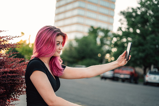 Unique Spunky Fashionable Young Woman with Fun Cute Bright Pink Dyed Hair Smiling and Using Her Mobile Cell Smartphone to Snap a Selfie to Stay Connected While on the Go Outdoors in the Summer