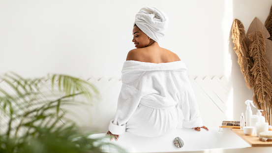 African American Lady Wearing White Bathrobe Sitting On Bathtub Back To Camera In Modern Bathroom At Home. Wellness And Spa, Female Beauty Routine Concept. Panorama, Rear View