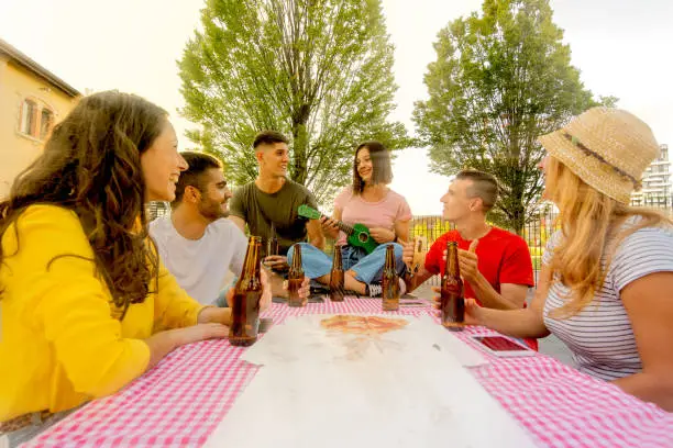 Photo of millennial people having fun with singing, eating pizza, drinking beers, young group of couples laughing around a table of a picnic, friendship and socialization after lockdown, warm filter effect