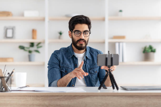 istock Arab man broadcasting from home, using smartphone and tripod, sitting at table and talking to phone camera 1322875076