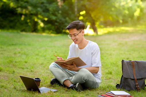 Outdoor Learning. Smiling Asian Male Student Reading Book On Lawn In Park, Studying And Preparing For Exams, Copy Space