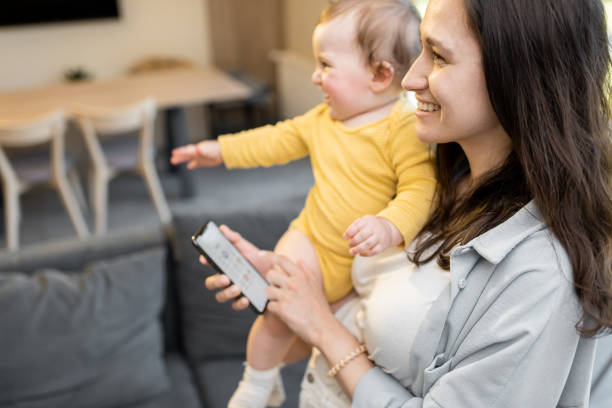 Mom controlling home devices by smartphone, standing with a baby at home Young mom controlling home devices by smartphone with running smart home application while nursing her baby at home. Concept of simple and easy management of a smart home smart home family stock pictures, royalty-free photos & images