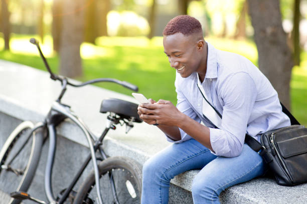 istock Joyful black man with modern bicycle using cellphone at urban park, copy space 1322875196