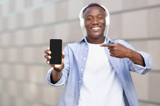 istock Handsome black man in headset pointing at cellphone with blank screen outdoors, mockup for mobile app or website design 1322876309