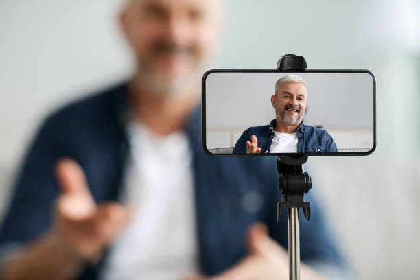 Mobile phone screen with cheerful senior man blogger Selective focus on mobile phone screen with cheerful senior man blogger broadcasting from home, blurred background, smartphone settled on tripod. Blogging, vlogging, video conference concept tripod stock pictures, royalty-free photos & images