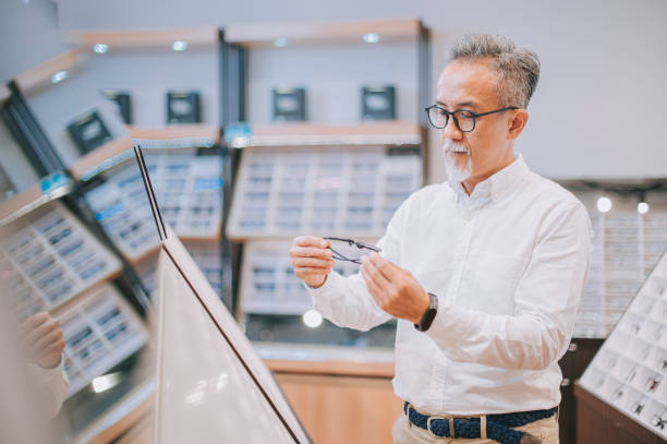 homem chinês sênior chinês experimentando óculos em frente ao espelho na loja de óptica - optometrist store retail glasses - fotografias e filmes do acervo