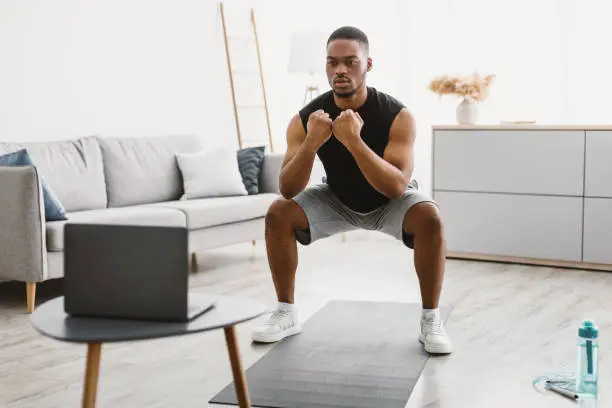 Domestic Workout. Athletic African American Guy Doing Deep Squats At Laptop During Online Training At Home. Male Fitness Routine. Sporty Black Man Exercising In Front Of Computer. Selective Focus