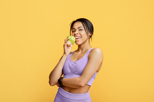 Black sporty lady with well fit body biting apple, eating fresh fruit after workout, standing ober yellow background, studio shot. Healthy food and fit body concept