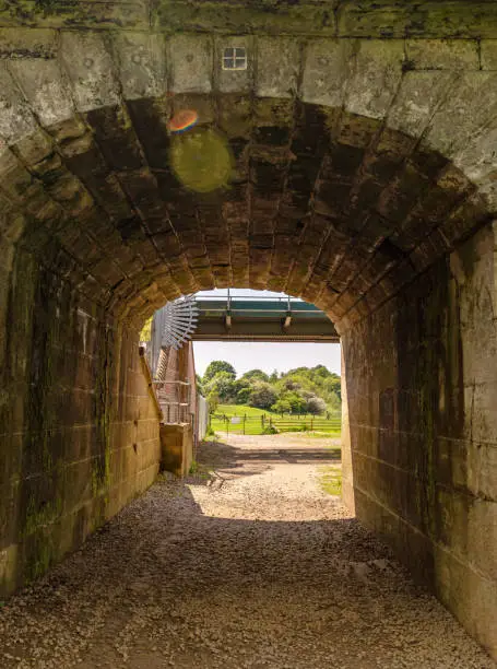 An old stone arch of a railway bridge over a pathway.  A metal bridge is adjacent and beyond is the open countryside.