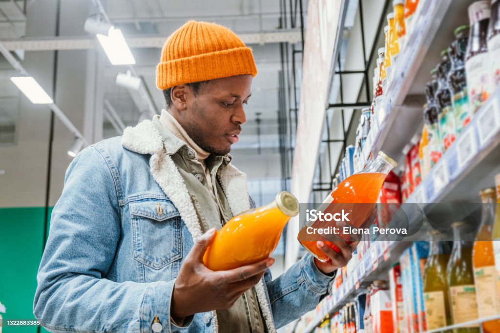 African man chooses natural juice in glass bottles in a grocery supermarket African man in a stylish orange hat and denim jacket chooses natural juice in glass bottles in a grocery supermarket standing at the shelves with products Shopping Stock Photo