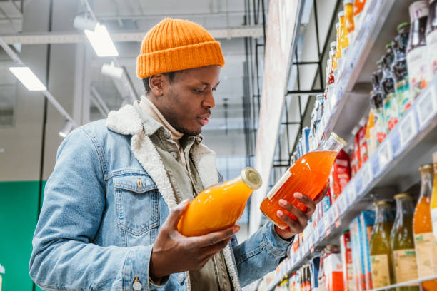 African man chooses natural juice in glass bottles in a grocery supermarket African man in a stylish orange hat and denim jacket chooses natural juice in glass bottles in a grocery supermarket standing at the shelves with products fruit juice stock pictures, royalty-free photos & images