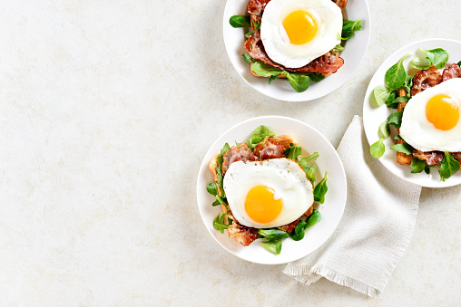 Open sandwiches with fried eggs, bacon and vegetable leaves on plate over light stone background with free text space. Top view, flat lay