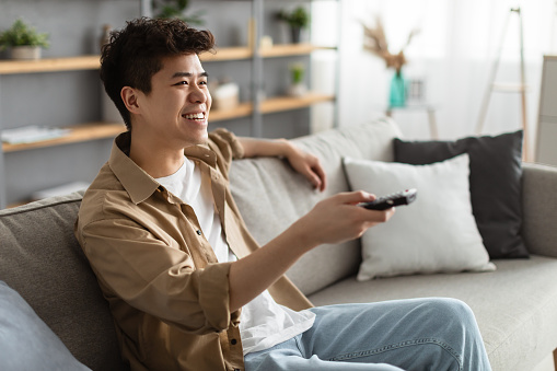 Leisure And Entertainment. Smiling asian guy watching TV show or film, holding remote control. Young man enjoying free time sitting on the comfortable couch at home in living room, free space