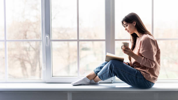 istock Woman sitting on windowsill drinking coffee reading book 1322886232