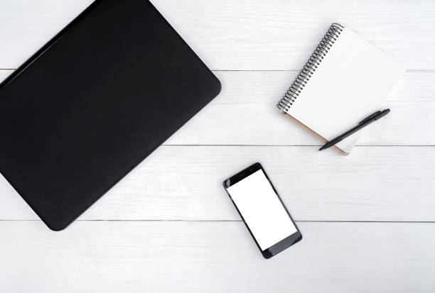 istock Top view on white wooden table with laptop computer, cell phone, and empty diary with pen, free space. Mobile phone with white screen, copy space 1322886261