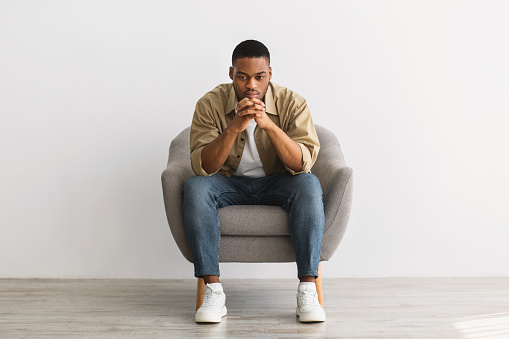 Thoughtful African American Man Thinking Sitting In Chair Over Gray Wall Background Indoors. Full-Length Shot Of Pensive Millennial Guy Reflecting On Problems, Front View