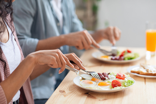 Unrecognizable couple using fork and knife while eating tasty breakfast in kitchen, cropped image of man and woman enjoying morning meal, having plate with fresh salad, eggs and sausages, closeup