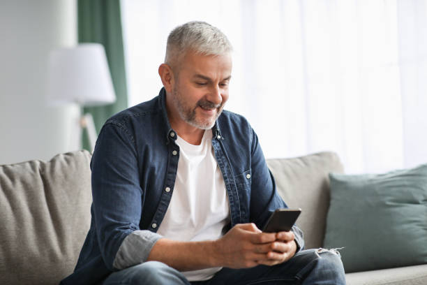 istock Smiling grey-haired mature man sitting on couch, using mobile phone 1322887081