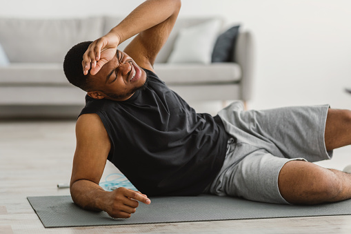 African American Guy Feeling Bad During Workout At Home, Man Suffering From Headache Having Hypertension Problem After Fitness Exercise Lying On Floor In Living Room. Sport And Healthcare