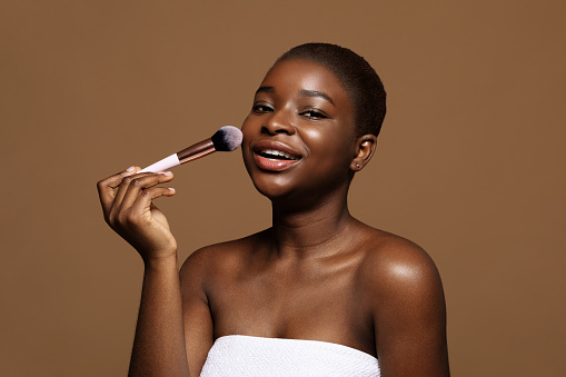 Daily Makeup Concept. Portrait of pretty black lady with clean skin holding make up brush, attractive african american female ready to apply blush tone, posing over brown background, free space