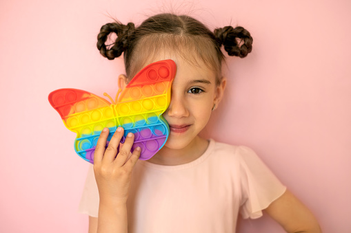 A beautiful cute little girl on a pink background covers one eye with a popit toy in the shape of a butterfly. Funny pigtails for a girl . Children's entertainment and educational pop it toys. simple dimple