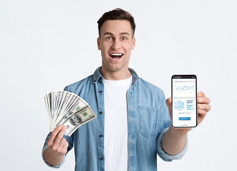 Excited guy holding dollar cash and smartphone with financial application on screen, recommending mobile app for trading online, standing isolated over white background, collage, copy space