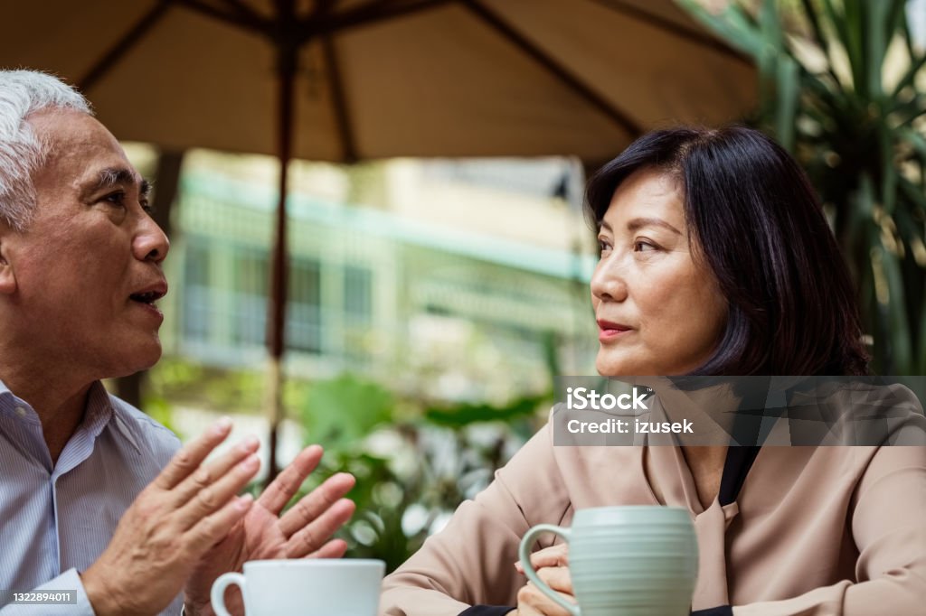 Senior couple talking in outdoor cafe Senior woman and man sitting at the table in small cafe, talking and drinking coffee. Asian couple enjoying time together. Sadness Stock Photo